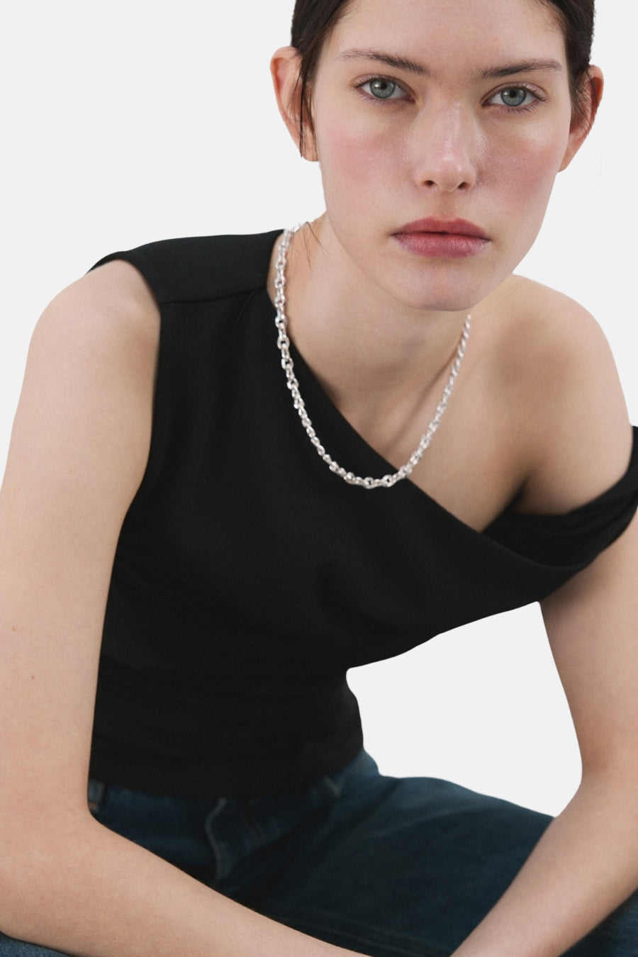 Haut noire sans manches de MANGO, avec une encolure asymétrique et portée avec une chaîne argentée et un jean.