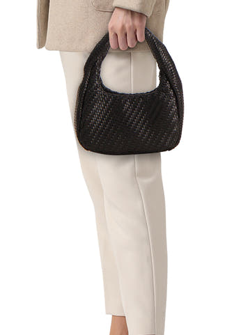 Sac à bandoulière noir de MANGO avec texture tressée, tenu par une personne portant une veste beige et un pantalon crème.