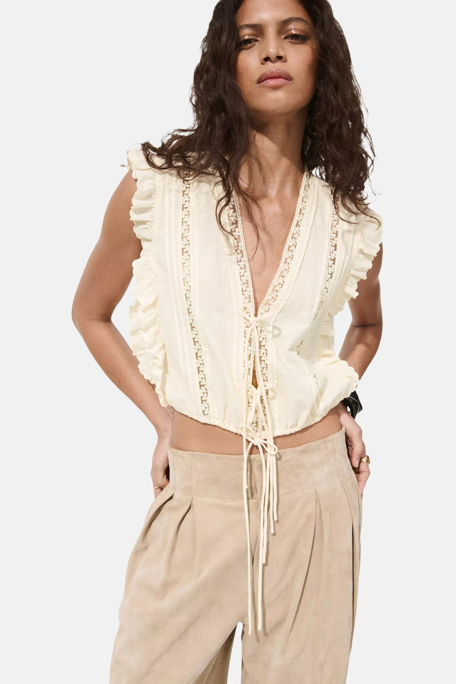 Beige mouwloze blouse met ruches en kant van MANGO, voorzien van strikdetail en diepe V-hals, gedragen met beige broek.