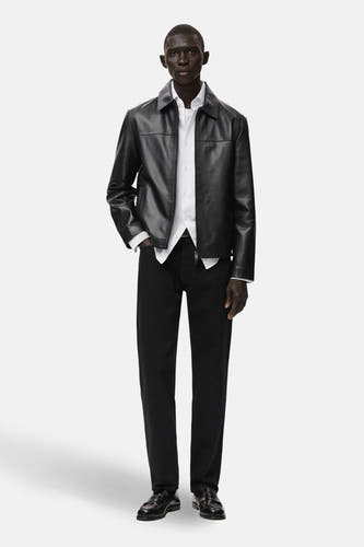 Manteau court noir de MANGO, avec des manches longues, porté sur une chemise blanche et un pantalon noir, complété par des chaussures noires.