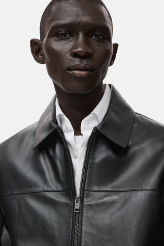 Manteau court noir de MANGO avec un col et une fermeture éclair, porté sur une chemise blanche.