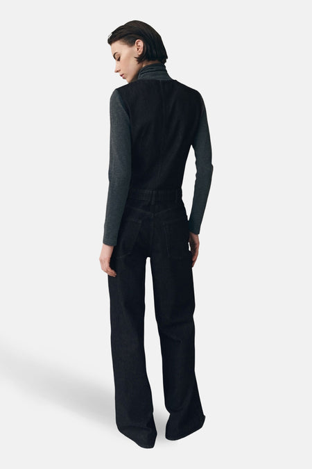 Donkerblauwe denim jumpsuit van MANGO, gezien van achteren, met lange, wijd uitlopende pijpen en een mouwloze top over een grijze coltrui.
