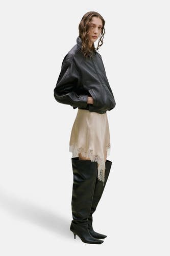 Jupe courte beige de MANGO, avec bordure en dentelle, combinée avec une veste en cuir et des bottes noires montantes.