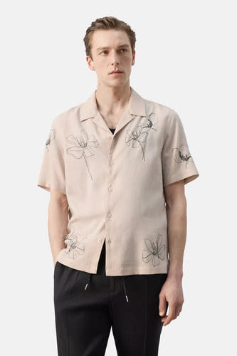 Chemise beige à manches courtes de MANGO, vue arrière, avec des fleurs brodées sur les manches, portée avec un pantalon noir.