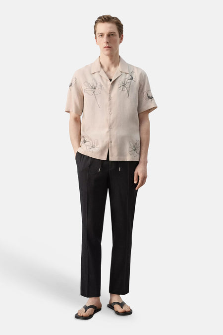 Chemise beige à manches courtes de MANGO, avec un motif floral noir, portée avec un pantalon noir et des tongs.