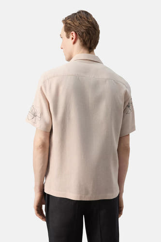 Beige overhemd met korte mouwen van MANGO, achterkant, met geborduurde bloemen op de mouwen, gedragen met een zwarte broek.