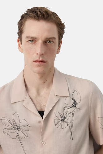 Chemise beige à manches courtes de MANGO, avec col ouvert, texture légère et broderies florales noires abstraites.
