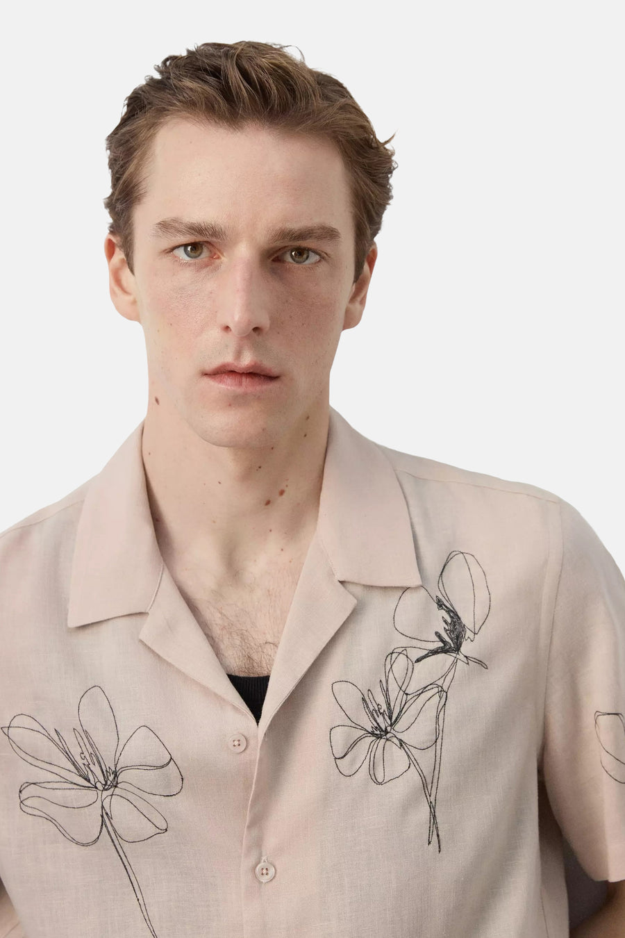 Chemise beige à manches courtes de MANGO, avec col ouvert, texture légère et broderies florales noires abstraites.