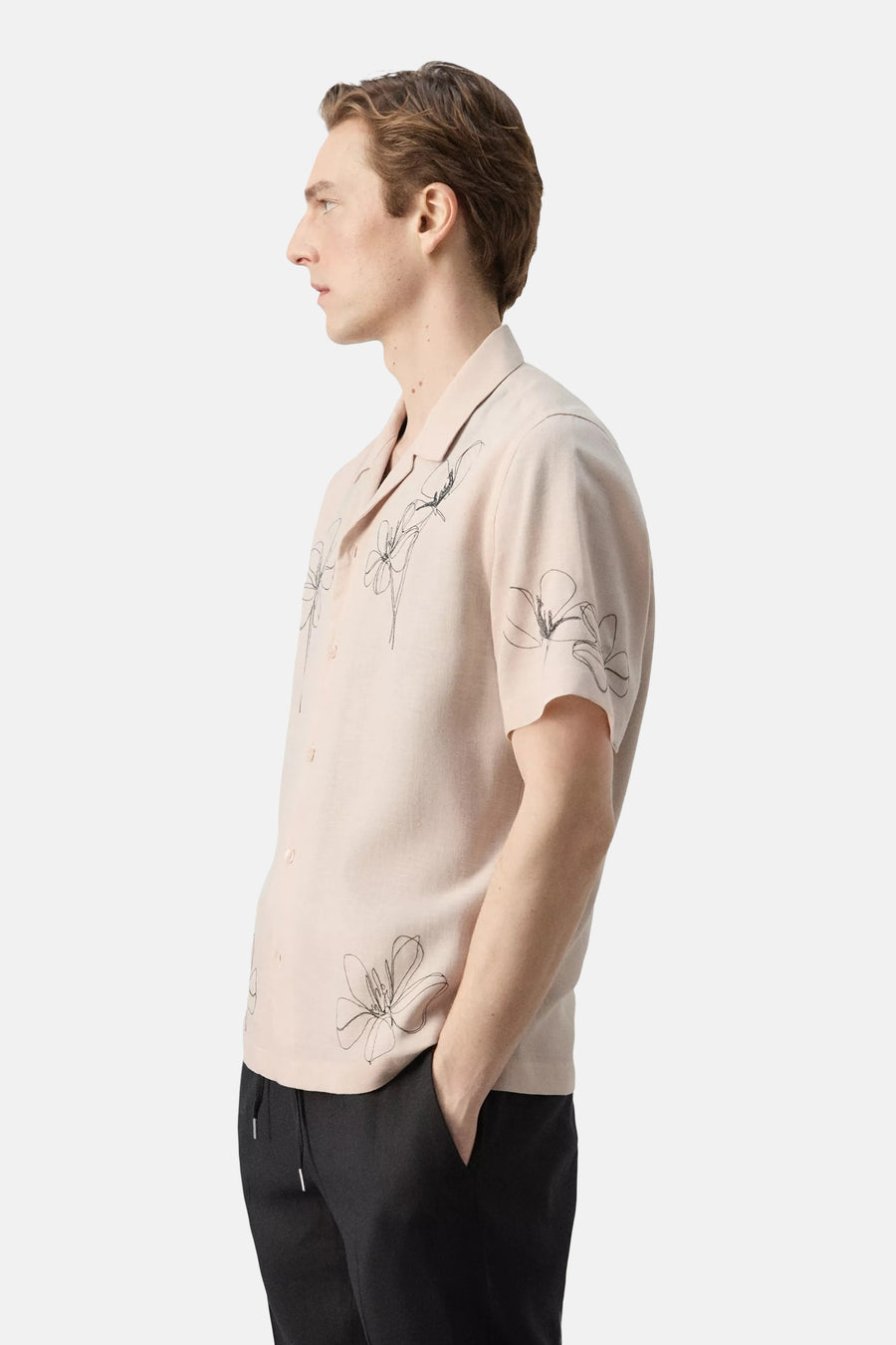 Vue de côté d'une chemise beige à manches courtes de MANGO, avec des motifs floraux noirs et un col cubain, portée par un homme en pantalon noir.