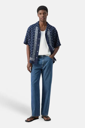 Chemise bleue à manches courtes de MANGO, avec motif cachemire et carreaux blancs, portée ouverte sur un débardeur blanc.