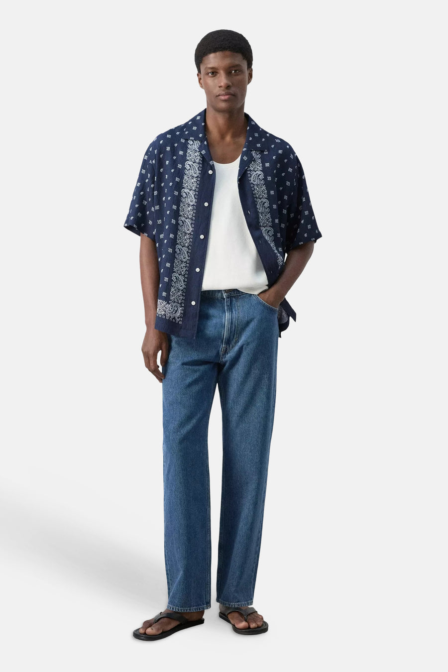 Chemise bleue à manches courtes de MANGO, avec motif cachemire et carreaux blancs, portée ouverte sur un débardeur blanc.