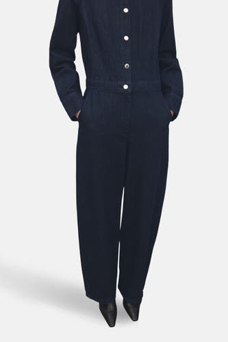 Donkerblauwe denim jumpsuit van MANGO, met lange mouwen, knoopsluiting en steekzakken, gecombineerd met zwarte schoenen.