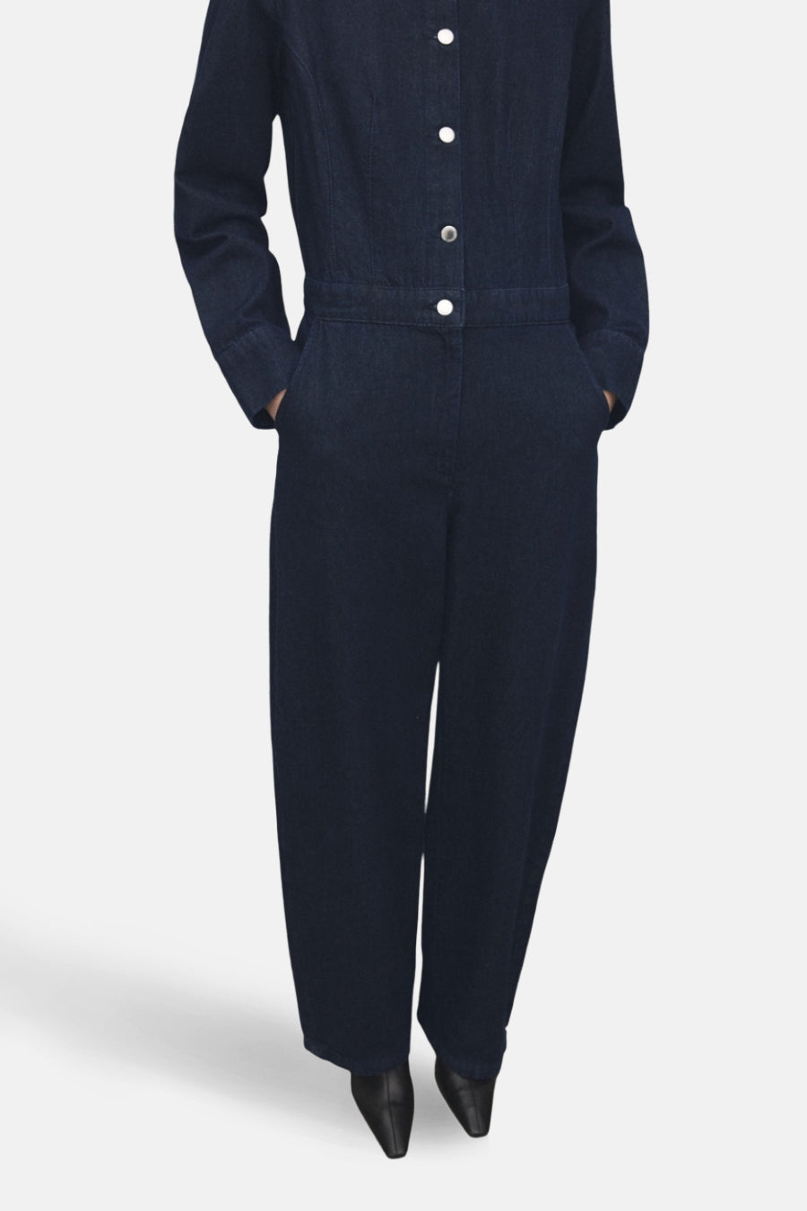Donkerblauwe denim jumpsuit van MANGO, met lange mouwen, knoopsluiting en steekzakken, gecombineerd met zwarte schoenen.