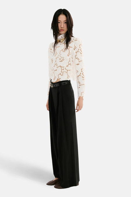 Chemisier blanc à manches longues de MANGO, avec motif floral et col montant, combiné avec un pantalon noir avec ceinture.