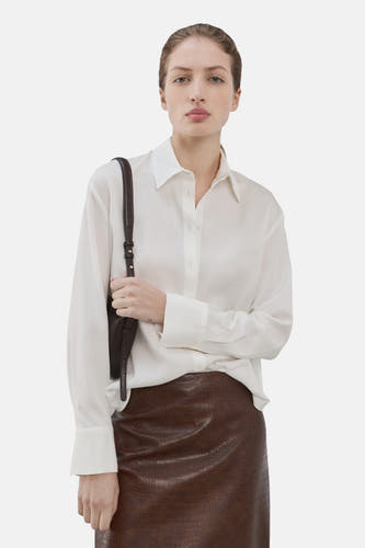Chemise blanche à manches longues de MANGO, vue de dos, associée à une jupe marron et un sac à bandoulière noir.