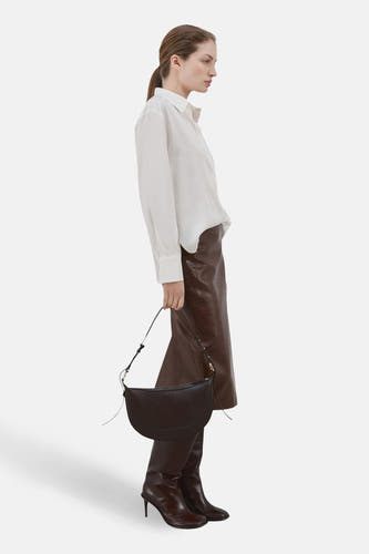 Chemise blanche à manches longues de MANGO, portée avec une jupe en cuir marron et des bottes à talons marron.