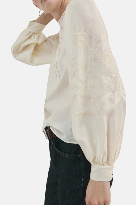 Blouse écru (manches longues) de MANGO, avec motifs floraux brodés sur les manches, combinée avec un jean foncé et une ceinture marron.