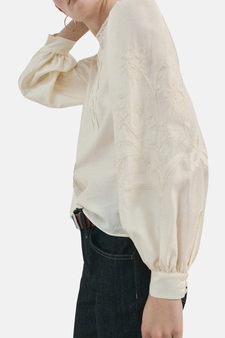 Blouse écru (manches longues) de MANGO, avec motifs floraux brodés sur les manches, combinée avec un jean foncé et une ceinture marron.