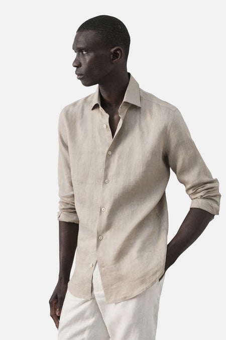 Chemise en lin beige de MANGO, à manches longues et fermeture boutonnée, portée avec un pantalon blanc.