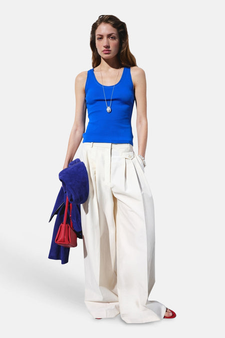 Débardeur côtelé bleu de MANGO, porté avec un pantalon blanc ample, une chaîne argentée, un sac à main rouge et une veste bleue.
