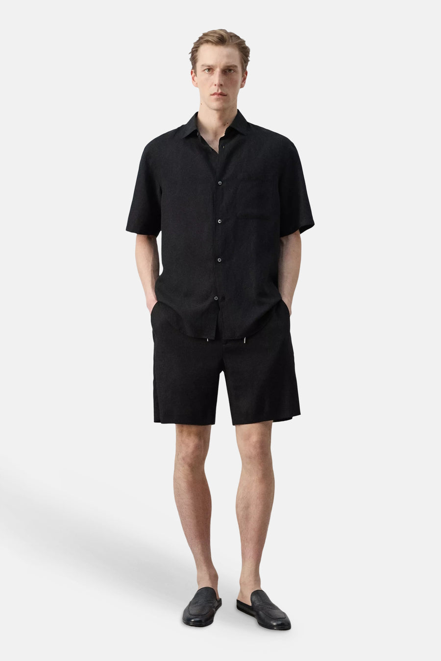 Chemise noire à manches courtes de MANGO, avec texture et poche poitrine, assortie à un short et des mules assortis.