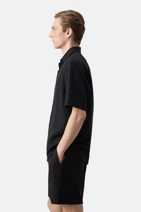 Chemise noire à manches courtes de MANGO, portée par un homme de profil, avec une main dans la poche de son short noir.