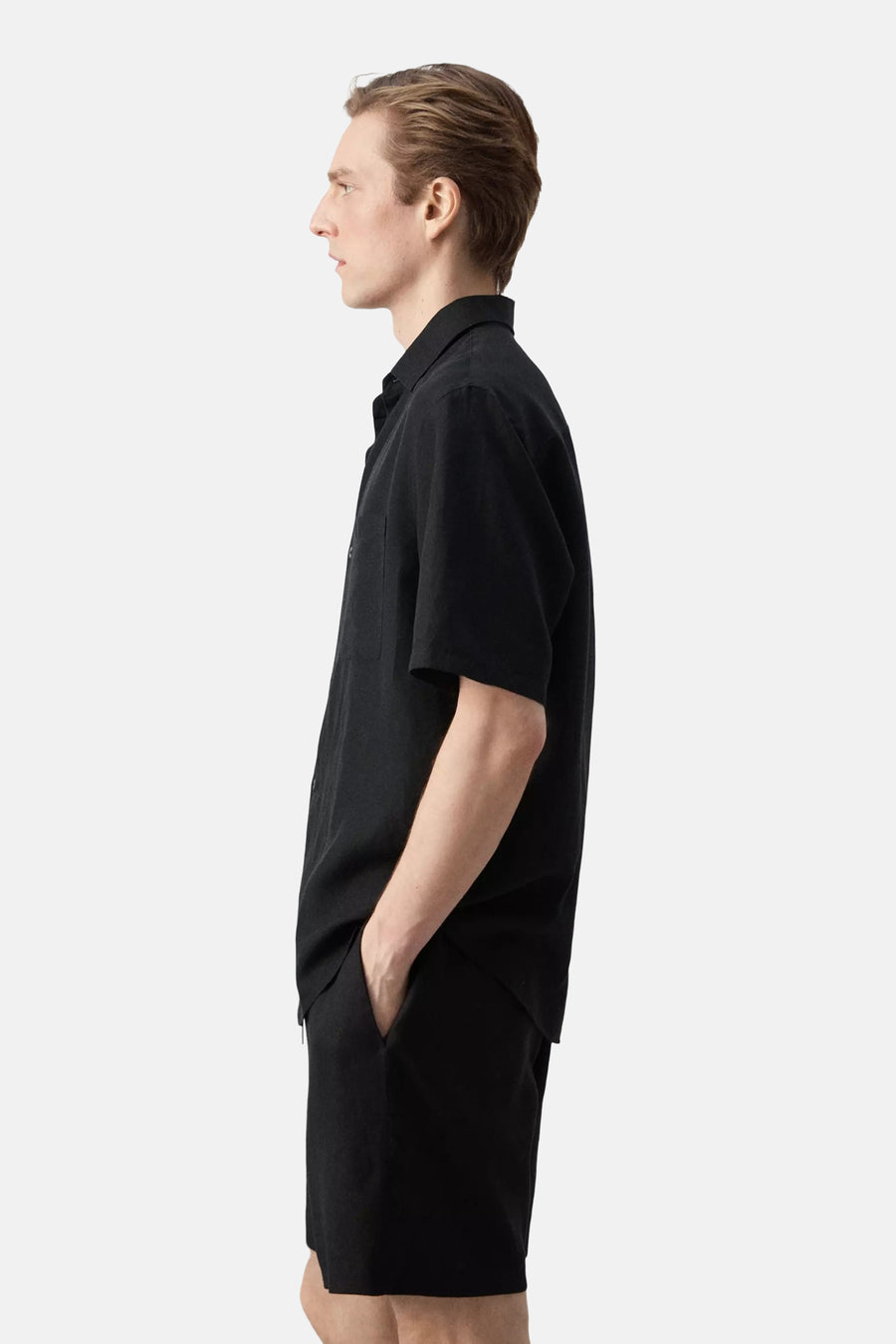 Chemise noire à manches courtes de MANGO, portée par un homme de profil, avec une main dans la poche de son short noir.