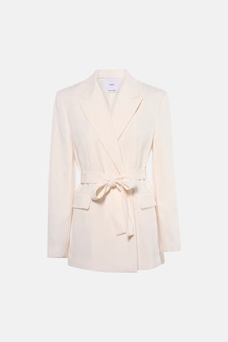 Blazer écru de MANGO, avec ceinture et porté avec un pantalon noir et des talons hauts.