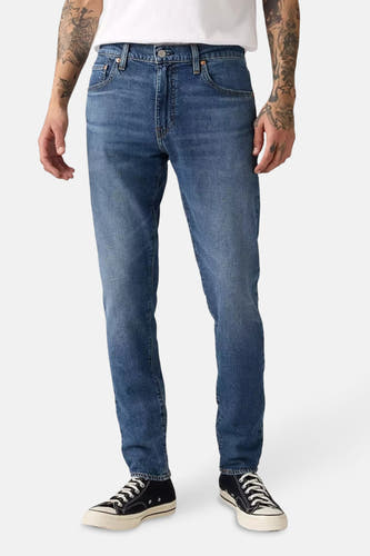 Jean tapered bleu Levi's porté avec des chaussures en toile noire et des tatouages visibles sur les bras.