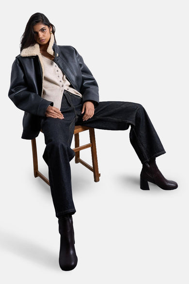 Veste écru de TOPSHOP, portée avec une veste en cuir noir à fourrure de mouton et des jeans bleu marine, assise sur une chaise en bois.