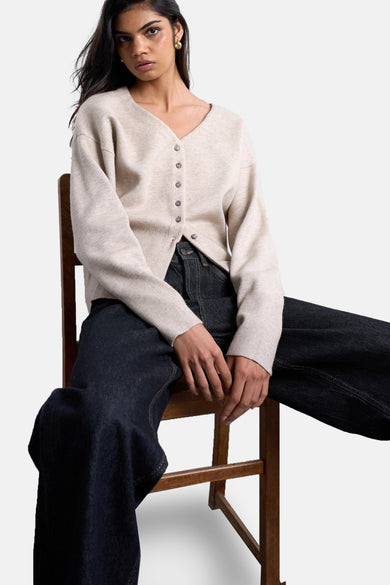 Cardigan écru de TOPSHOP, porté avec des jeans foncés, assis sur une chaise en bois.