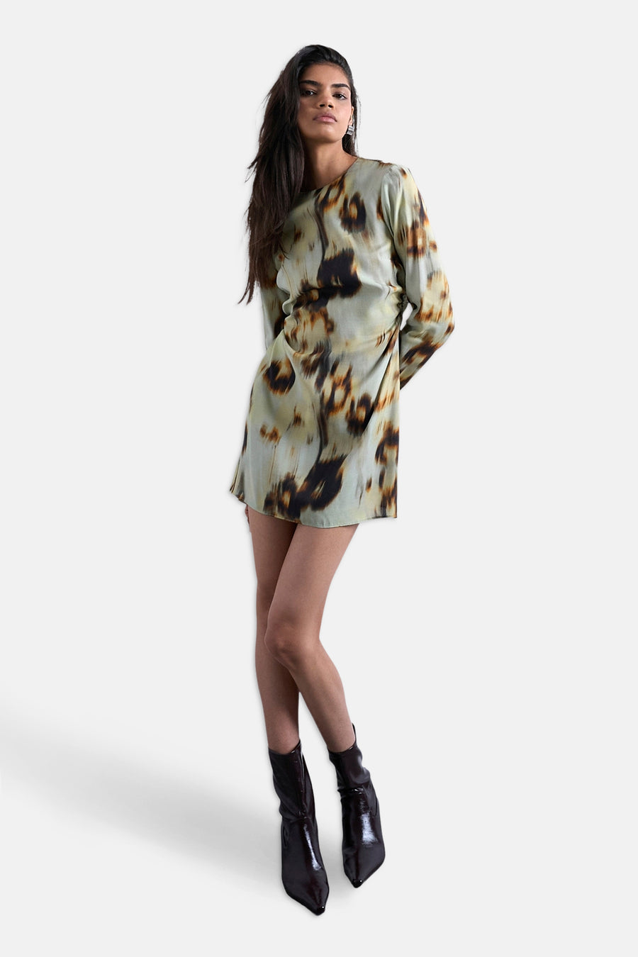 Vrouw in bedrukte jurk en zwarte laarzen, staand voor witte achtergrond.
