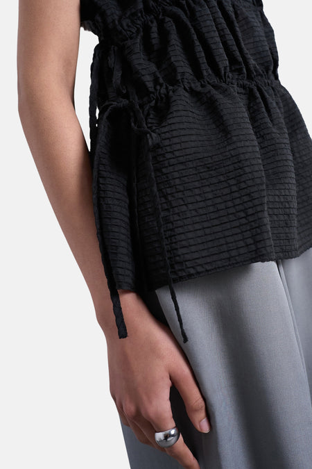 T-shirt noir de TOPSHOP, avec une texture plissée, fermeture à cordon sur le côté, associé à un pantalon gris et une bague argentée.