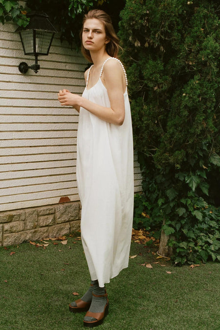 Wit mouwloos kleedje van MANGO met parelbeslag aan de schouderbandjes, lichte stof, lifestyle shot in tuin, model draagt grijze sokken en bruine sandalen