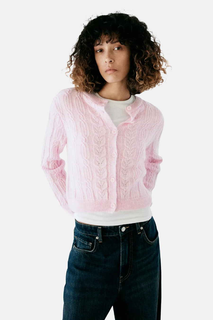 Roze gebreide gilet met kabelpatroon van MANGO, voorzien van knopen, gedragen over een witte top en donkerblauwe jeans, front view.