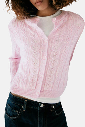 Roze gebreide gilet met kabelpatroon en knopen van MANGO, gedragen over een witte trui en donkerblauwe jeans, close-up voorzijde.