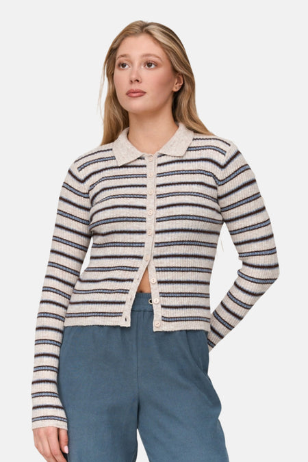 Een vrouw met blond haar draagt een gestreepte cardigan en blauwe broek.