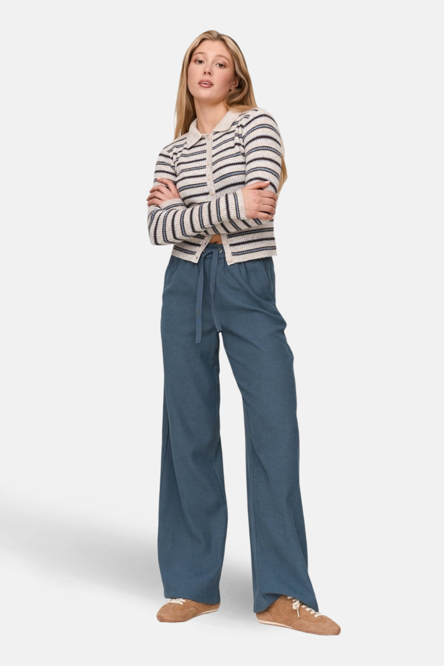 Vrouw met gestreepte cardigan, blauwe broek en bruine schoenen, staand met gekruiste armen.