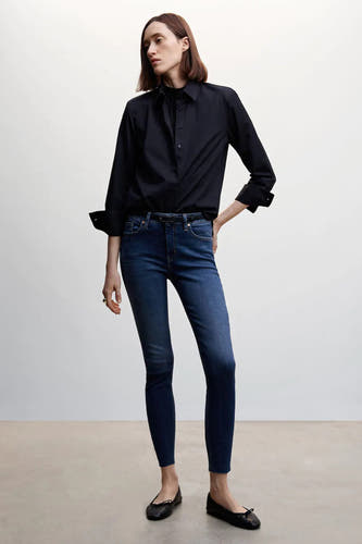Jean skinny bleu de MANGO avec ourlets bruts, stylisé avec un chemisier noir et des ballerines.