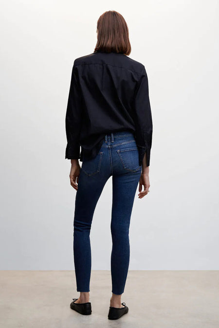 Jean skinny bleu moyen en denim de MANGO avec ourlet effiloché, vu sur un mannequin de dos, associé à une chemise noire et des ballerines noires.