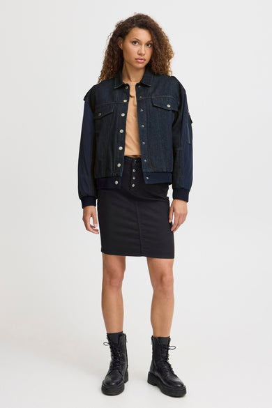 Veste en jeans bleu marine de PULZ, avec des manches longues bleu marine et des boutons argentés, portée avec une jupe noire et des bottes noires.