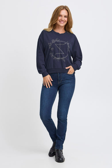 Pull bleu de PULZ, avec une impression décorative sur la devanture, associé à des jeans et des bottes noires.