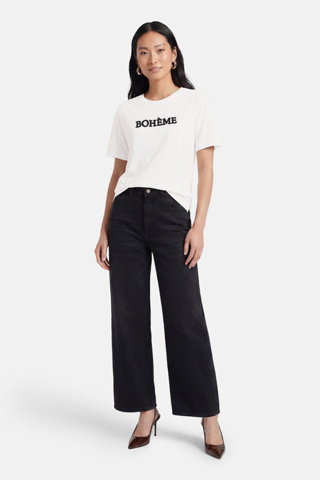 T-shirt blanc de PULZ avec "BOHÈME" en noir sur le devant, porté avec un jean noir à jambes larges et des talons pointus marron.