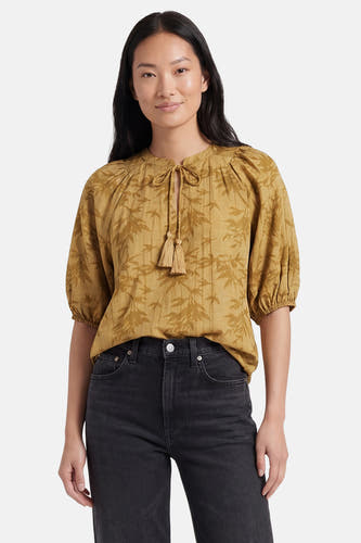 Chemisier jaune à manches courtes de PULZ, avec imprimé feuilles, fermeture à nouer avec pompons et manches bouffantes, porté sur un jean noir.