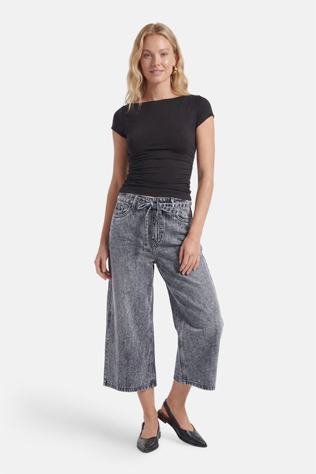 Bermuda en denim gris de PULZ avec taille haute, jambes larges et ceinture à nouer, porté avec un t-shirt noir et des chaussures plates noires.
