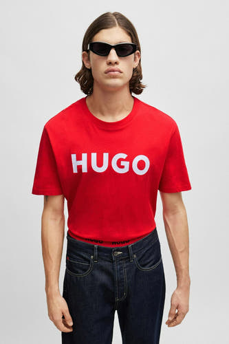 T-shirt rouge HUGO, avec un logo blanc sur la poitrine, porté avec un jeans foncé et des lunettes de soleil.