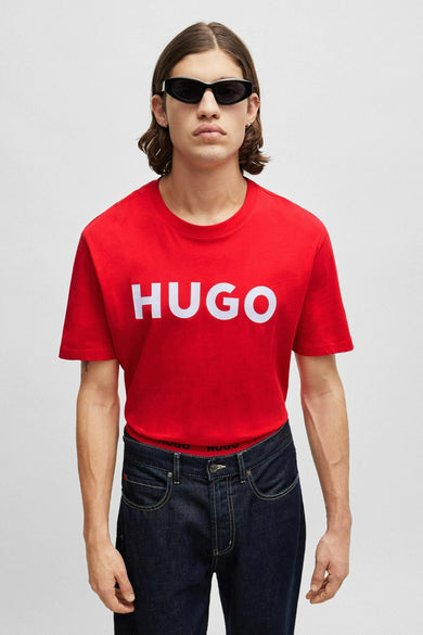 T-shirt rouge HUGO, avec un logo blanc sur la poitrine, porté avec un jeans foncé et des lunettes de soleil.