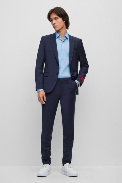 Man in blauw pak met lichtblauw overhemd en witte sneakers, staand.