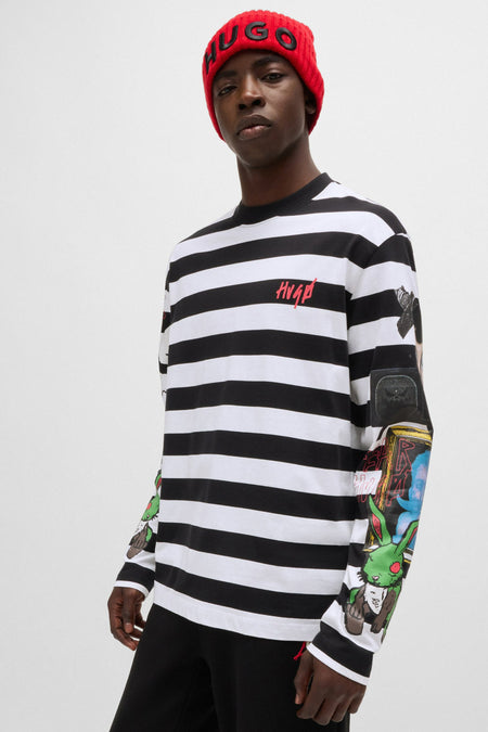 T-shirt à manches longues rayé noir et blanc de HUGO, avec logo rouge sur la poitrine et imprimés colorés sur les manches, porté par un mannequin avec un bonnet rouge.
