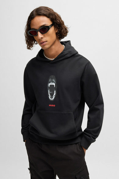 Man met zonnebril en zwarte HUGO hoodie met hondentandenprint, parelketting en zwarte broek.
